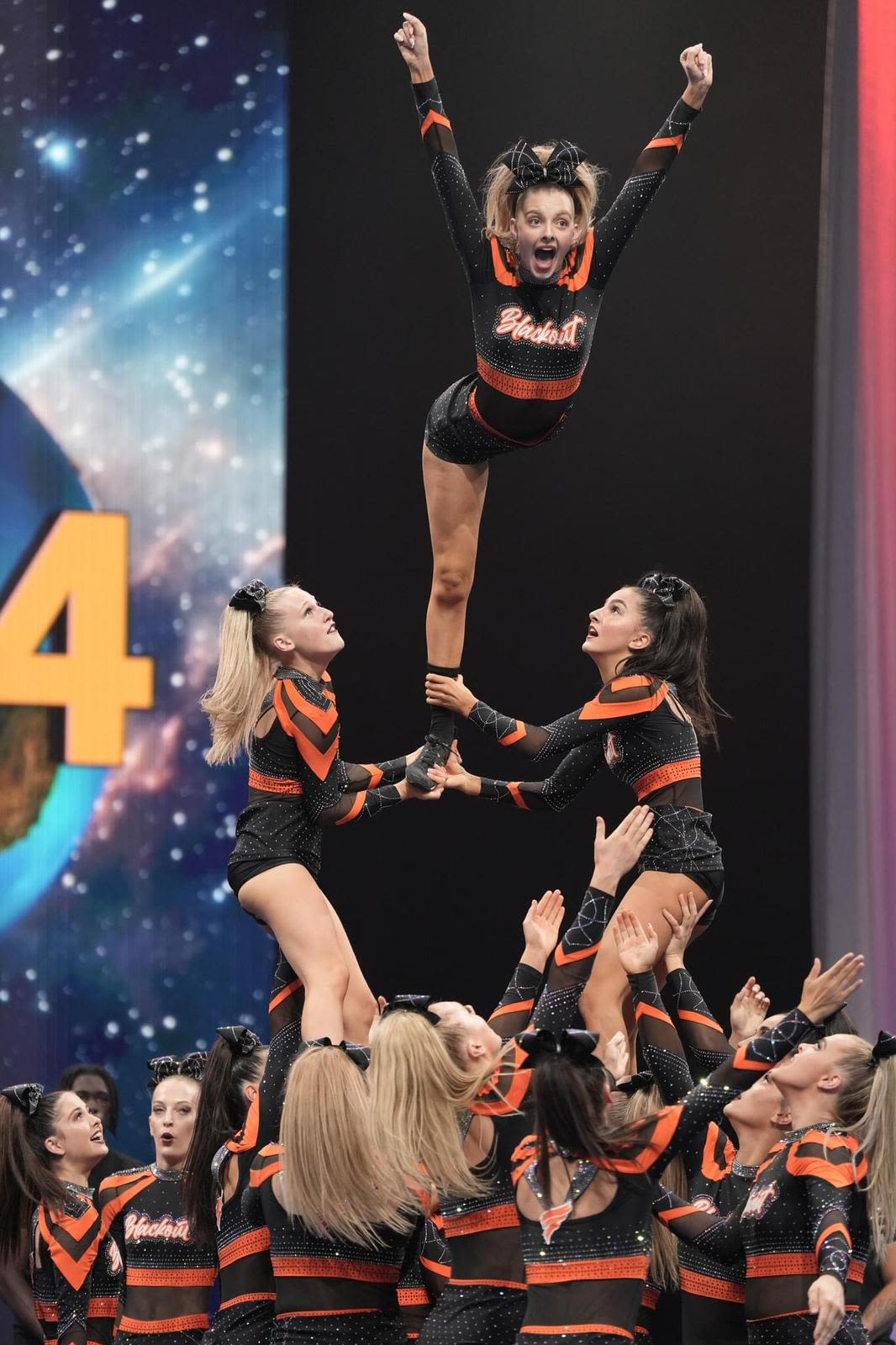 Two girls in orange and black outfits, one brunette and one blonde, are lifted into the air. They are holding a blonde girl up, who has her arms raised above her head and is in motion.